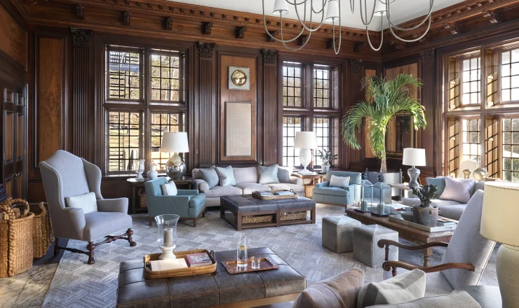 Luxurious living room with dark wood paneling, oversized windows, and a grand chandelier overhead.