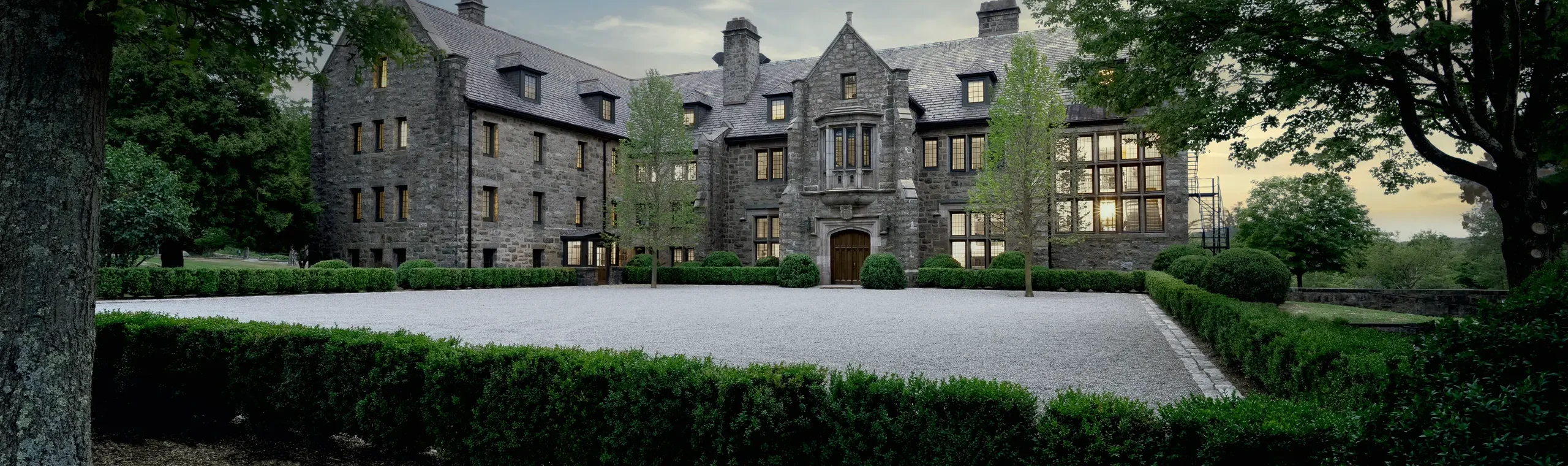 Stone manor house with a steep slate roof, central arched entrance, and lit windows, set in a manicured gravel courtyard framed by trimmed hedges and mature trees.