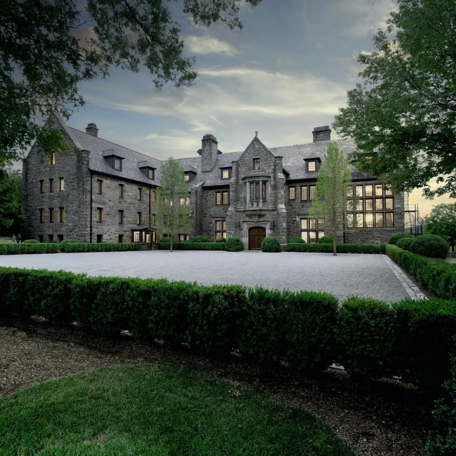 Stone manor house with a steep slate roof, central arched entrance, and lit windows, set in a manicured gravel courtyard framed by trimmed hedges and mature trees.