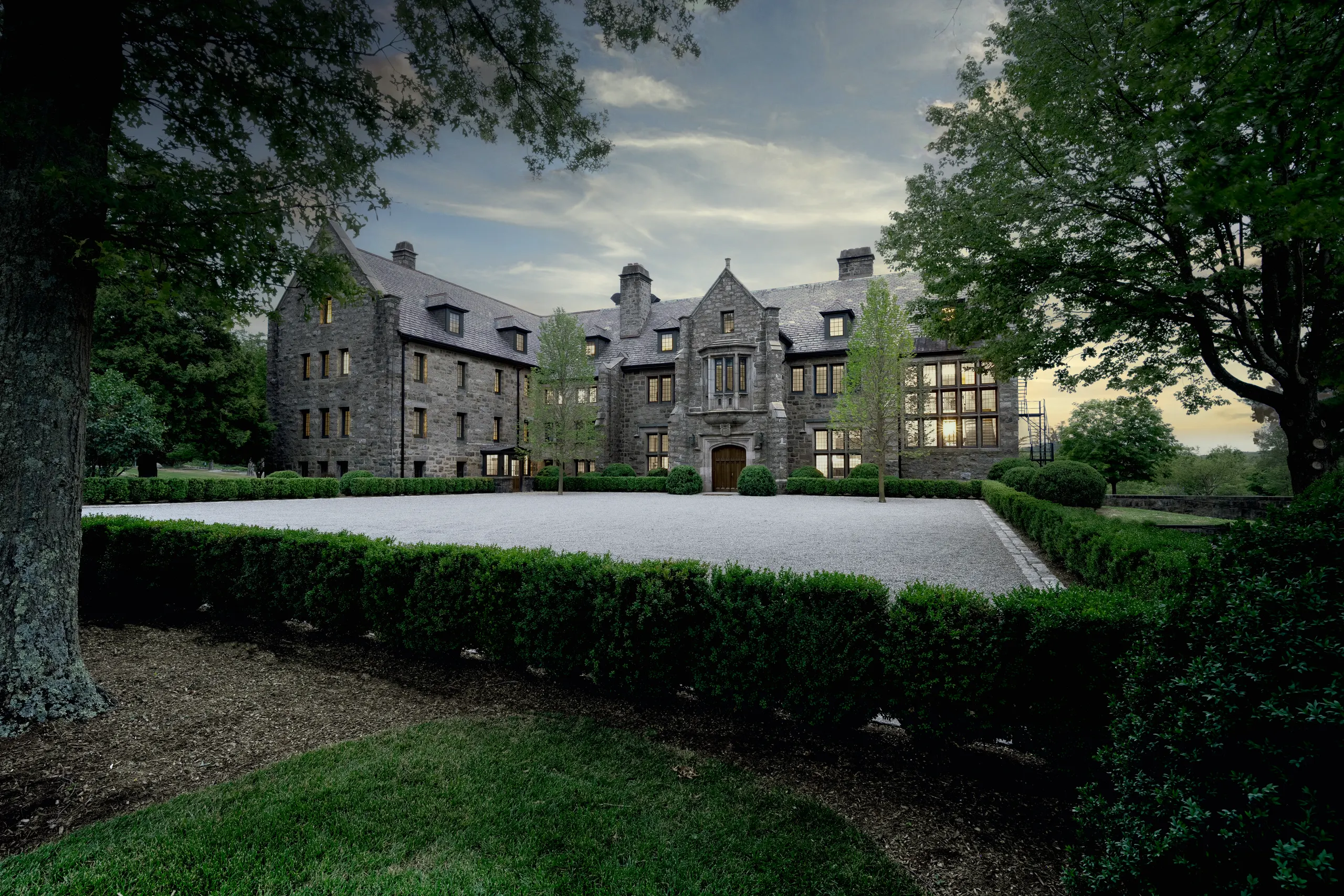 Stone manor house with a steep slate roof, central arched entrance, and lit windows, set in a manicured gravel courtyard framed by trimmed hedges and mature trees.