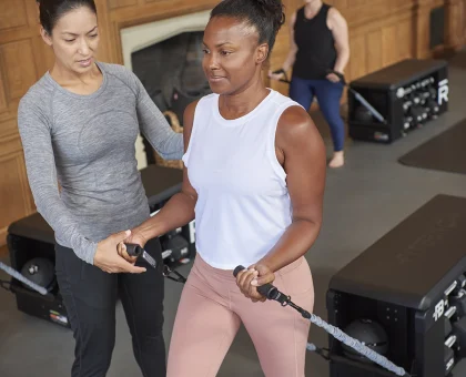 Instructor guides a woman in a white tank top using resistance bands during a workout in a wood-paneled gym; another person watches in the background in workout gear.