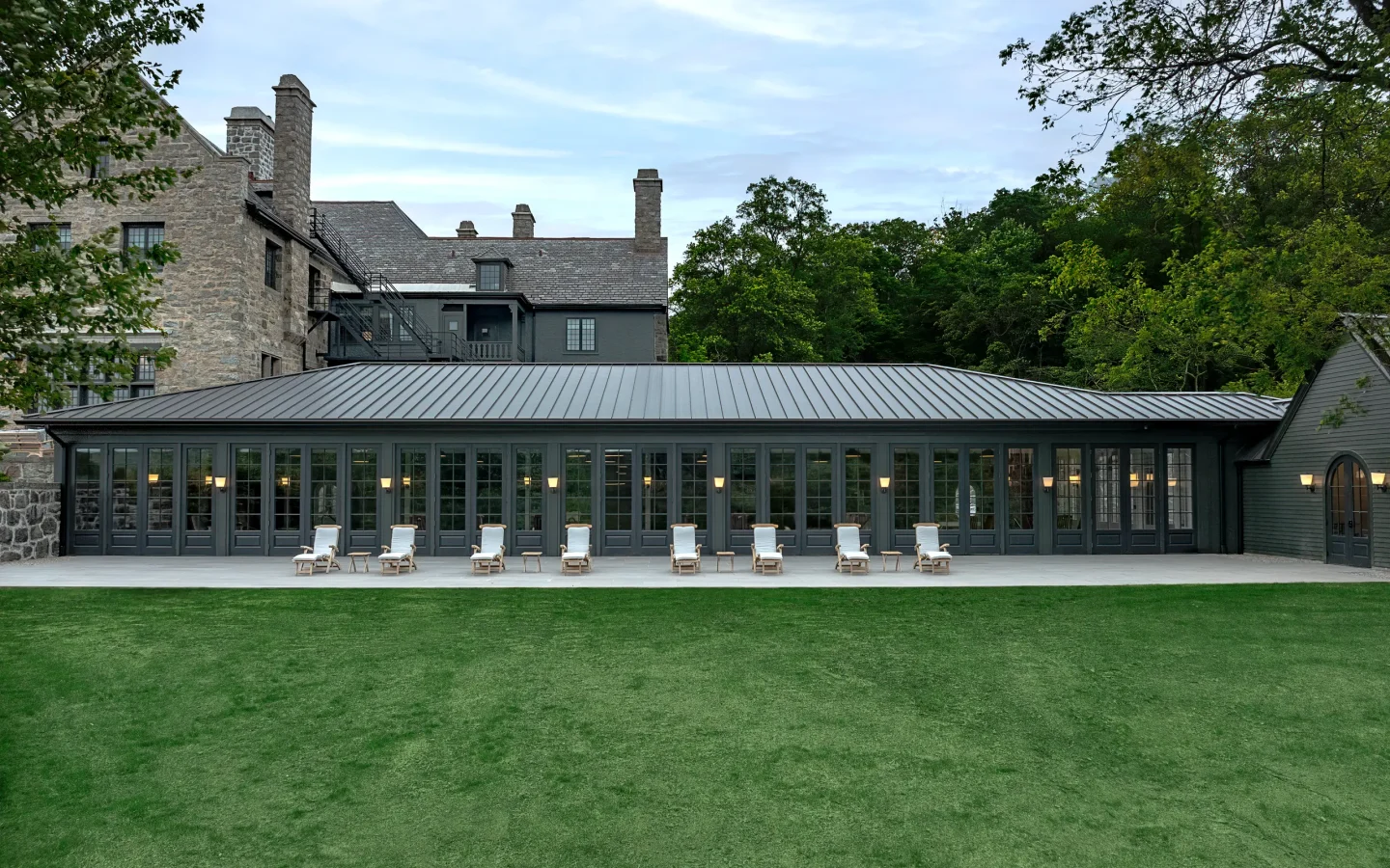 Stone building with slate roof and a row of glass-fronted doors, outdoor lounge chairs lined up on a covered terrace overlooking a green lawn.
