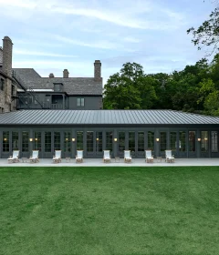 Stone building with slate roof and a row of glass-fronted doors, outdoor lounge chairs lined up on a covered terrace overlooking a green lawn.