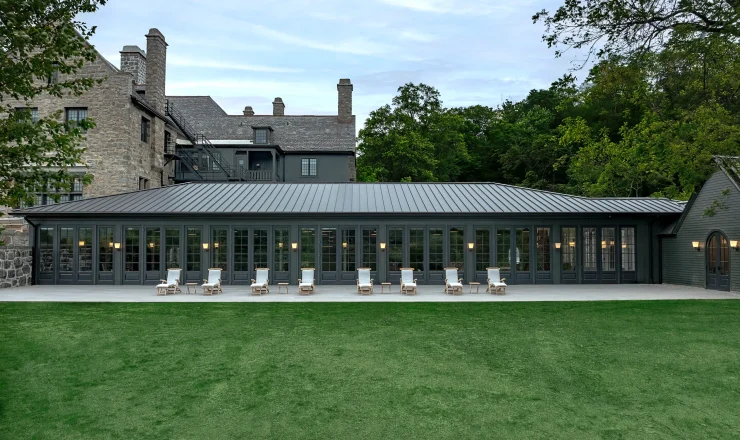 Stone building with slate roof and a row of glass-fronted doors, outdoor lounge chairs lined up on a covered terrace overlooking a green lawn.