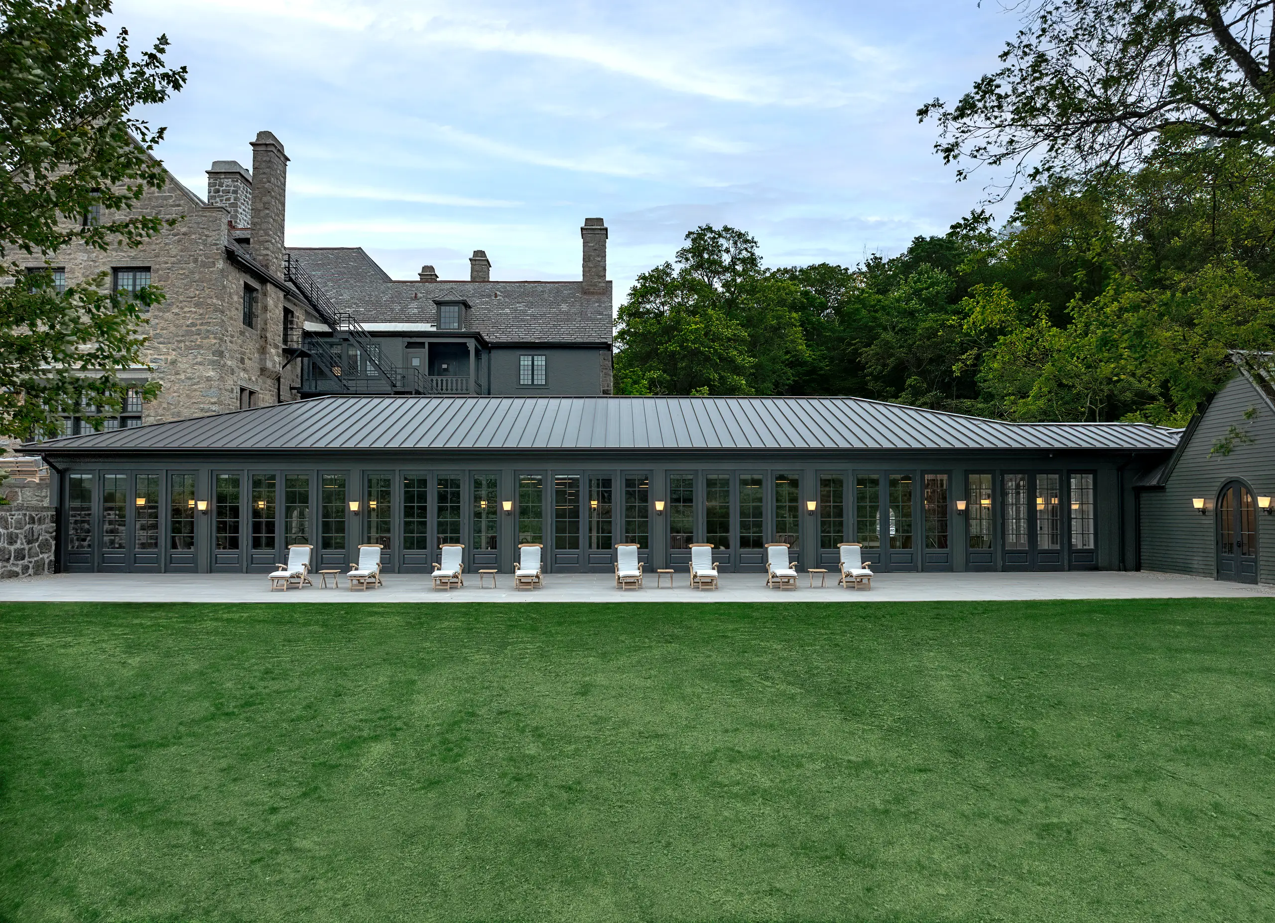 Stone building with slate roof and a row of glass-fronted doors, outdoor lounge chairs lined up on a covered terrace overlooking a green lawn.