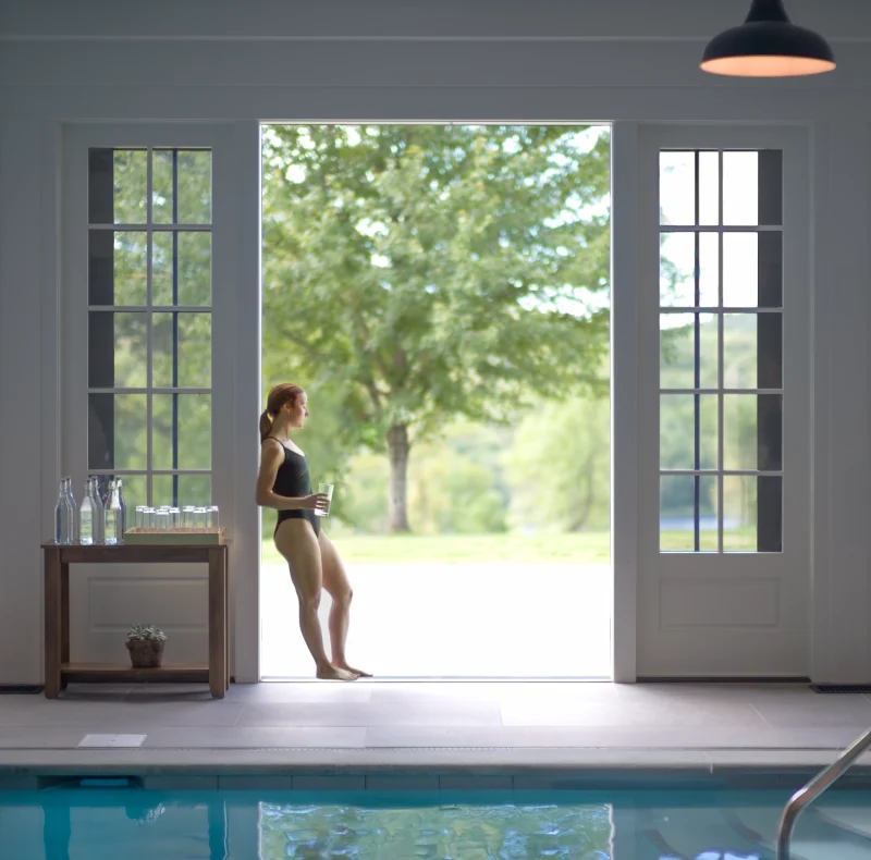 Woman in a black swimsuit stands in the doorway of an indoor pool, holding a glass and looking outside at greenery.
