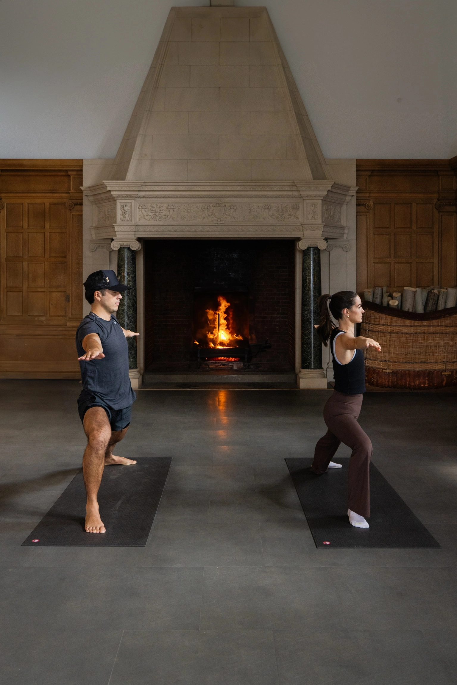 Two people practice yoga on dark mats in a room with a large ornate fireplace, facing opposite directions.