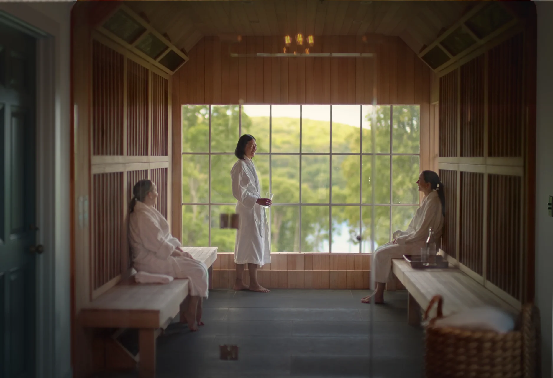 Inside a wooden sauna or spa room, four people in white robes sit or stand along benches, near a large window with greenery outside.