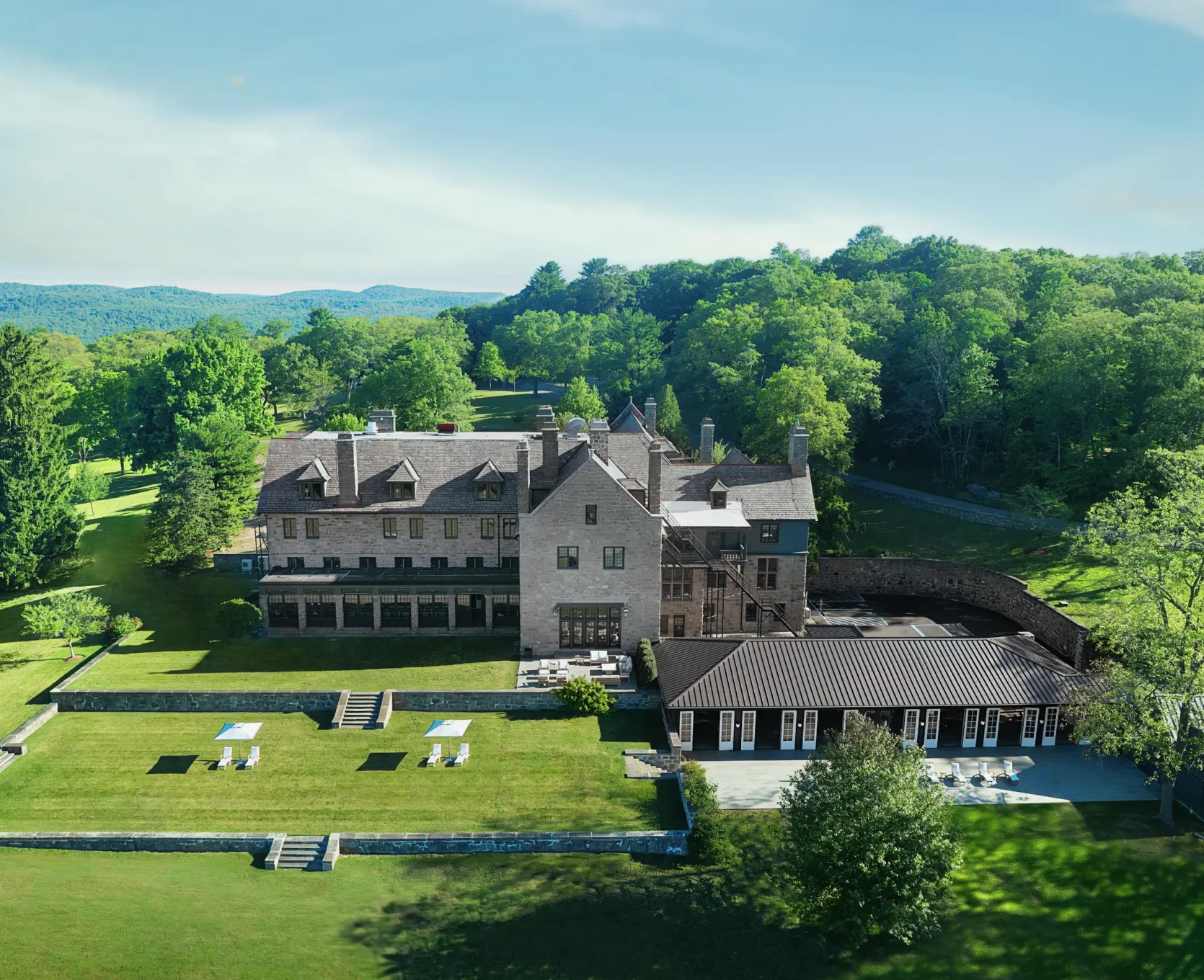 Aerial view of a stone campus building with a green lawn, outdoor seating, and surrounding trees with forested hills in the distance.