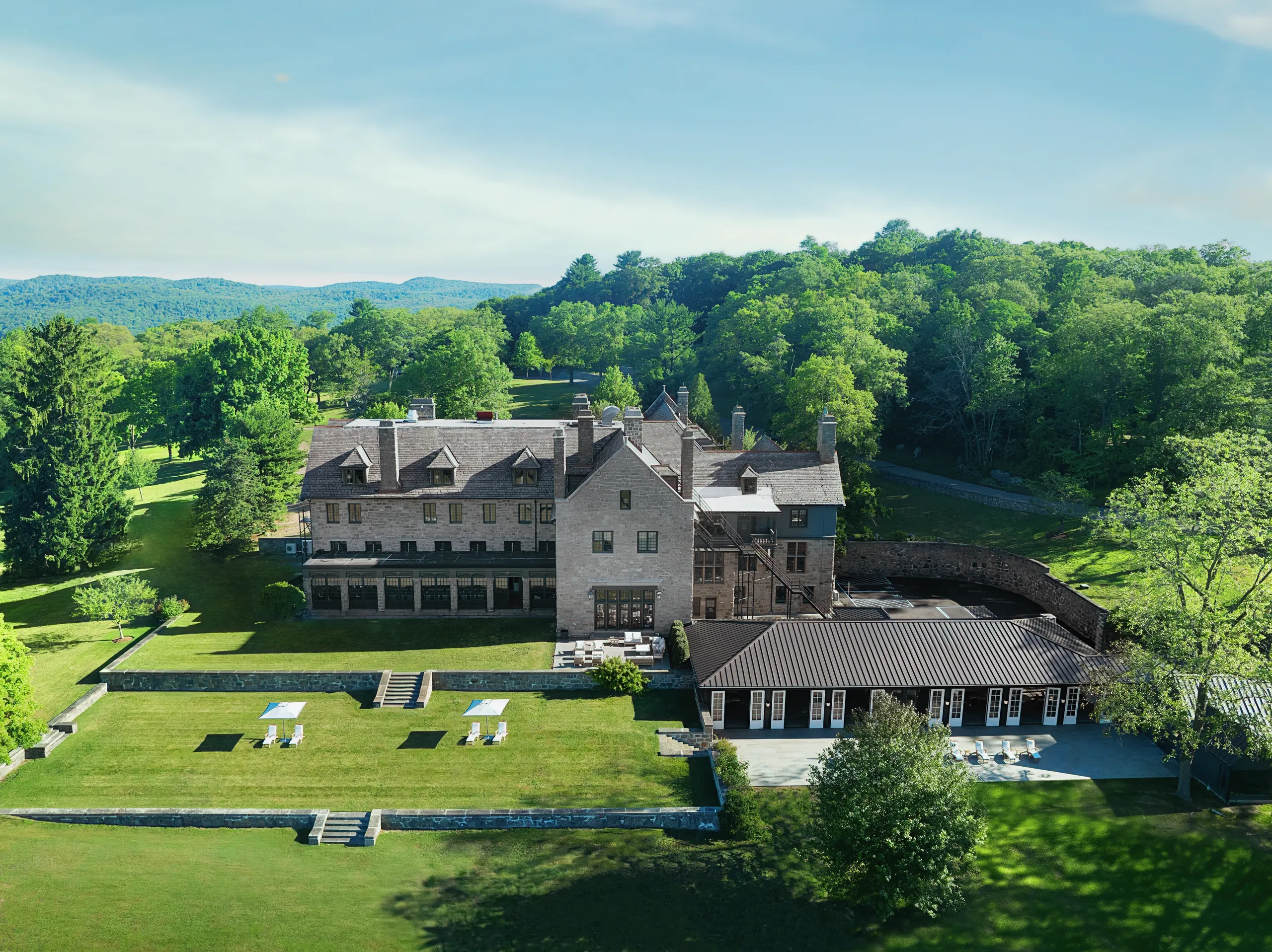 Aerial view of a stone campus building with a green lawn, outdoor seating, and surrounding trees with forested hills in the distance.