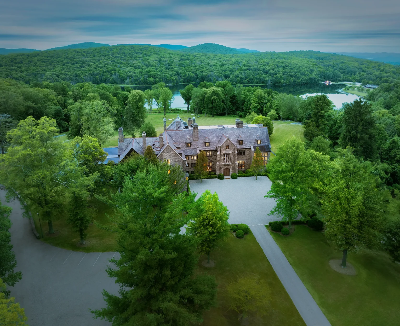 Stone manor surrounded by trees with a lake and forested hills in the distance under a cloudy sky.