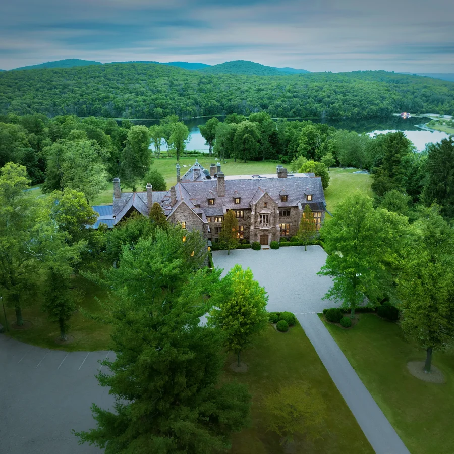 Stone manor surrounded by trees with a lake and forested hills in the distance under a cloudy sky.
