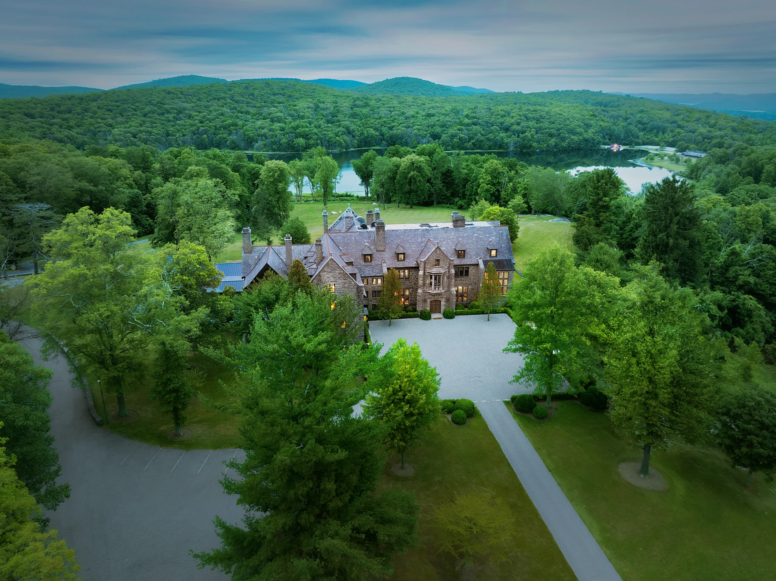 Stone manor surrounded by trees with a lake and forested hills in the distance under a cloudy sky.