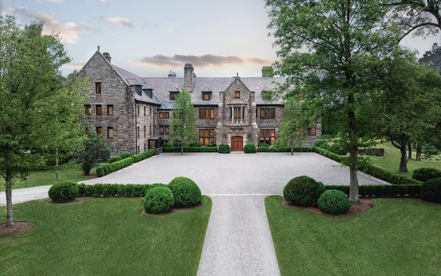 Front view of a large stone mansion with a gravel courtyard and neatly trimmed evergreen shrubs, surrounded by trees.