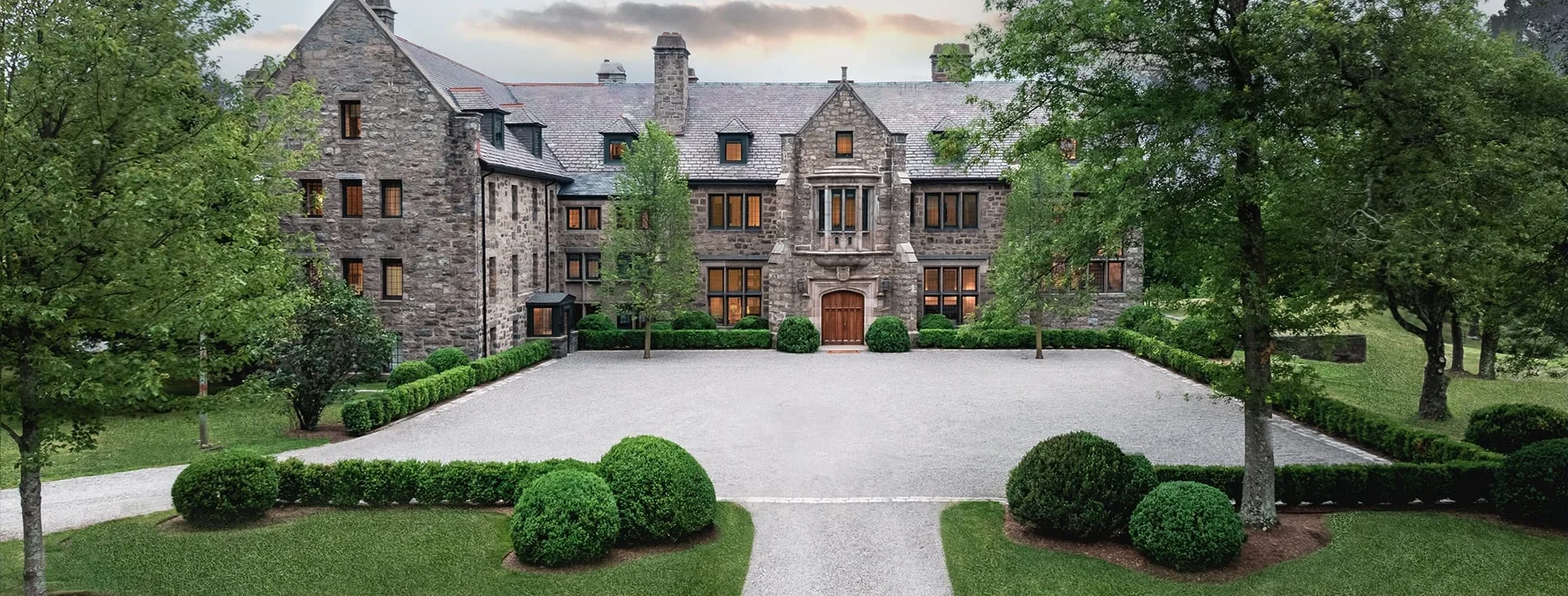 Front view of a large stone mansion with a gravel courtyard and neatly trimmed evergreen shrubs, surrounded by trees.