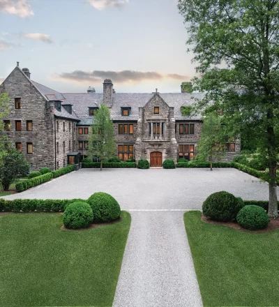 Front view of a large stone mansion with a gravel courtyard and neatly trimmed evergreen shrubs, surrounded by trees.