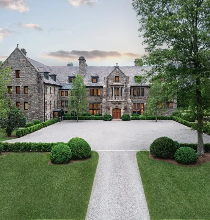 Front view of a large stone mansion with a gravel courtyard and neatly trimmed evergreen shrubs, surrounded by trees.