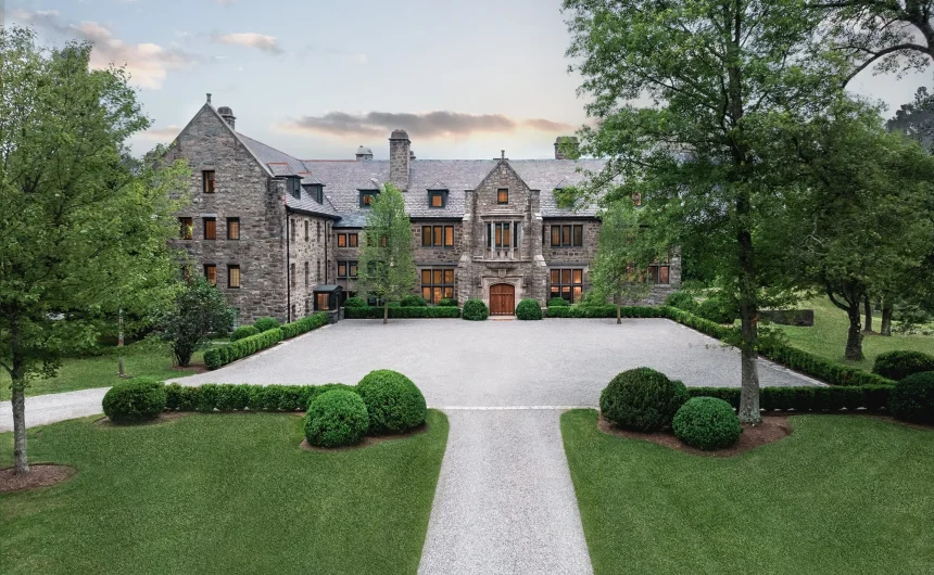 Front view of a large stone mansion with a gravel courtyard and neatly trimmed evergreen shrubs, surrounded by trees.