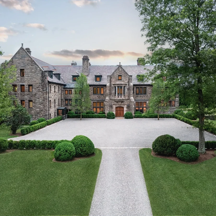 Front view of a large stone mansion with a gravel courtyard and neatly trimmed evergreen shrubs, surrounded by trees.