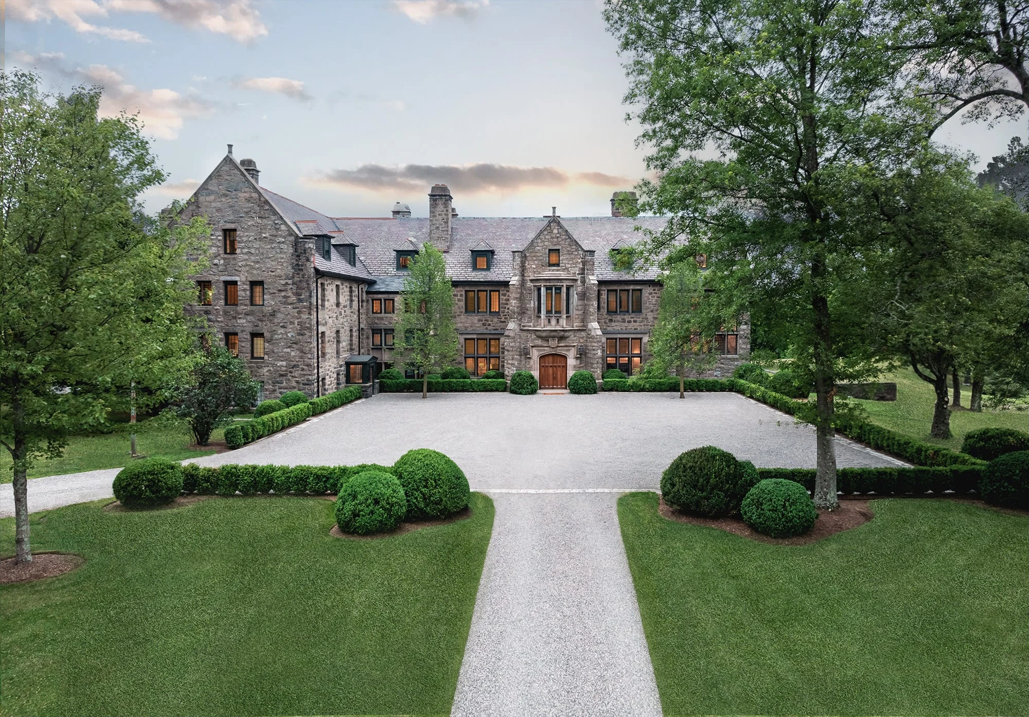 Front view of a large stone mansion with a gravel courtyard and neatly trimmed evergreen shrubs, surrounded by trees.