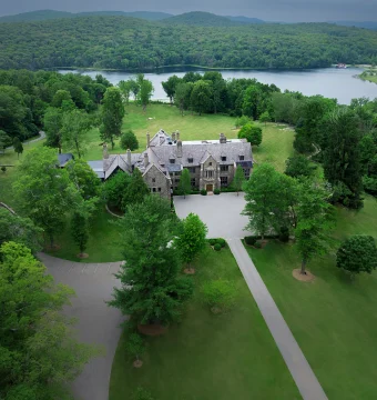 Aerial view of a stone mansion with a gray roof surrounded by a manicured lawn and dense trees, with a lake and hills in the distance.