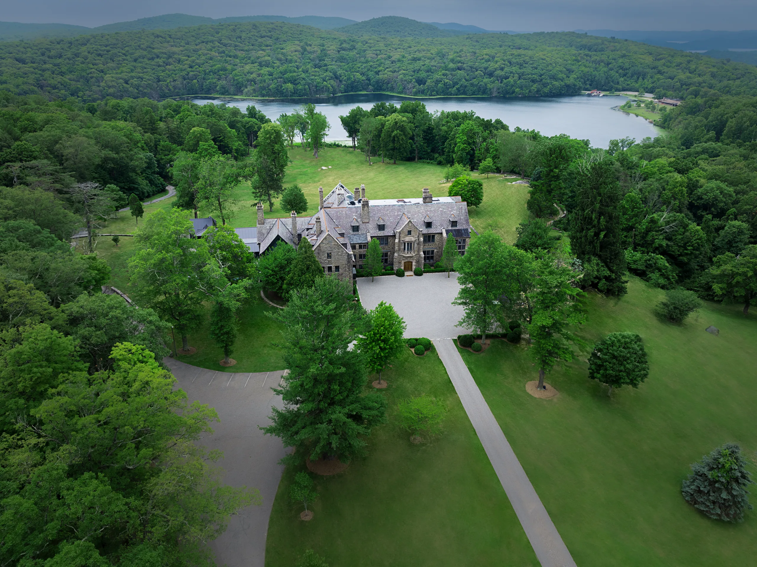 Aerial view of a stone mansion with a gray roof surrounded by a manicured lawn and dense trees, with a lake and hills in the distance.