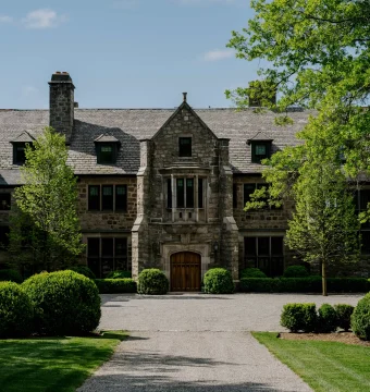 Historic stone mansion with a central arched wooden doorway, flanked by symmetrical windows and manicured shrubs on a gravel driveway, under a blue sky with trees overhead.