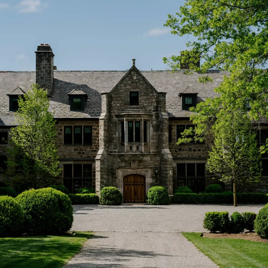 Historic stone mansion with a central arched wooden doorway, flanked by symmetrical windows and manicured shrubs on a gravel driveway, under a blue sky with trees overhead.