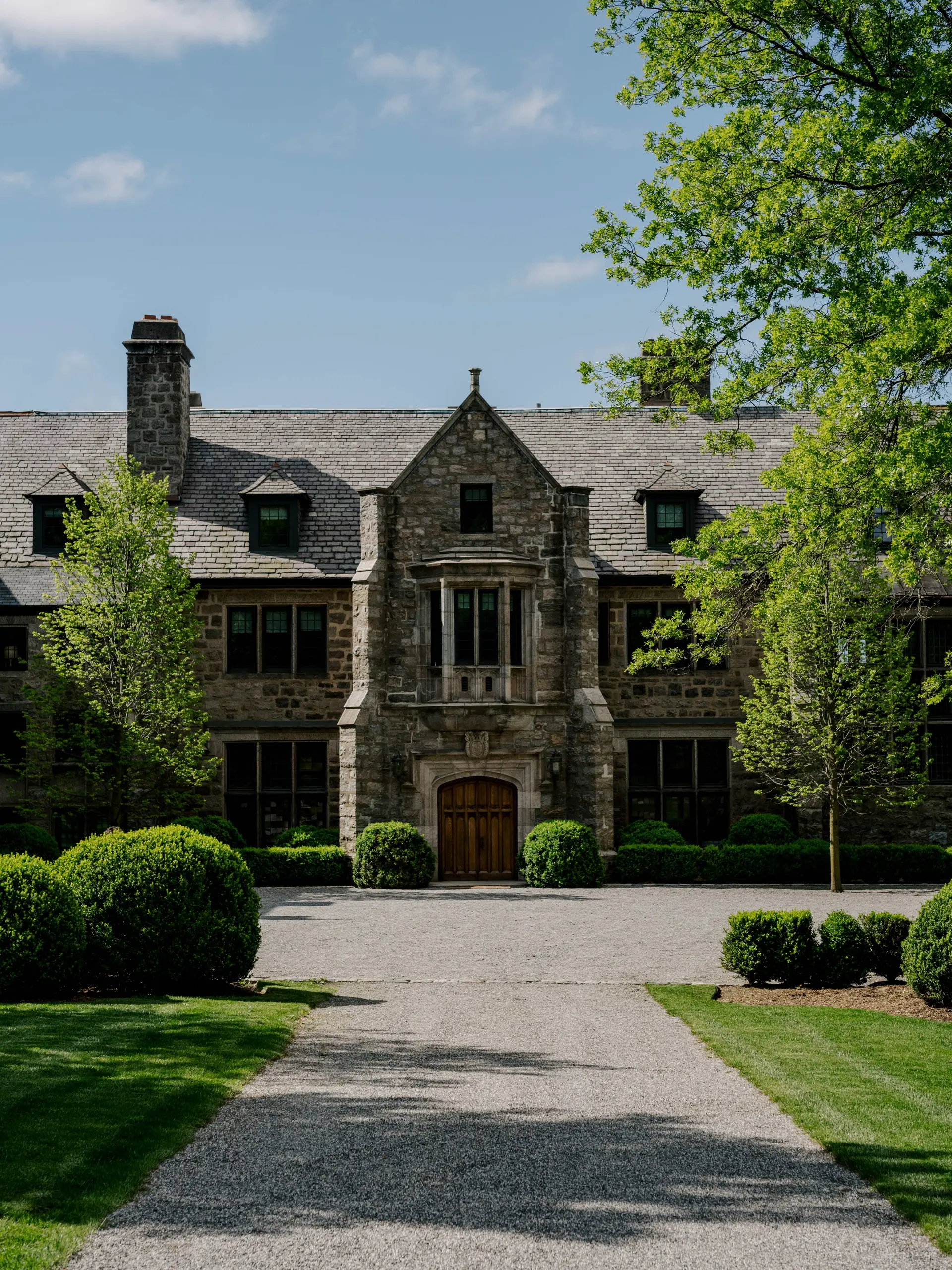 Historic stone mansion with a central arched wooden doorway, flanked by symmetrical windows and manicured shrubs on a gravel driveway, under a blue sky with trees overhead.