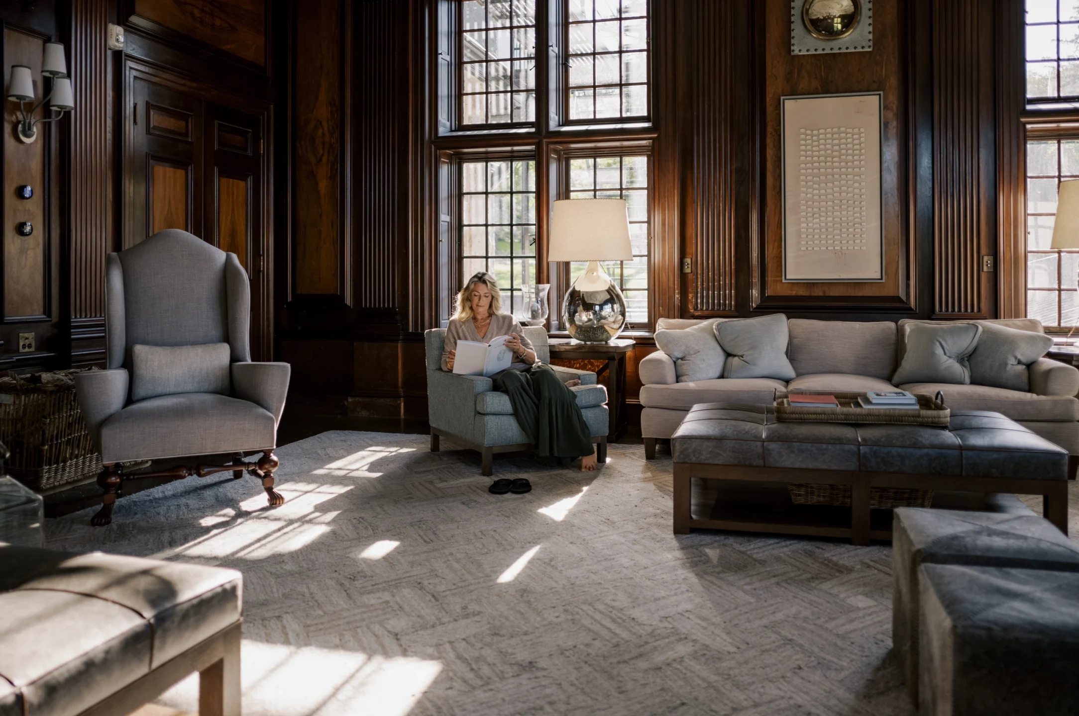 Woman reading a book in a blue armchair in a dark wood-paneled living room with large grid windows and a glass-lamp table nearby.