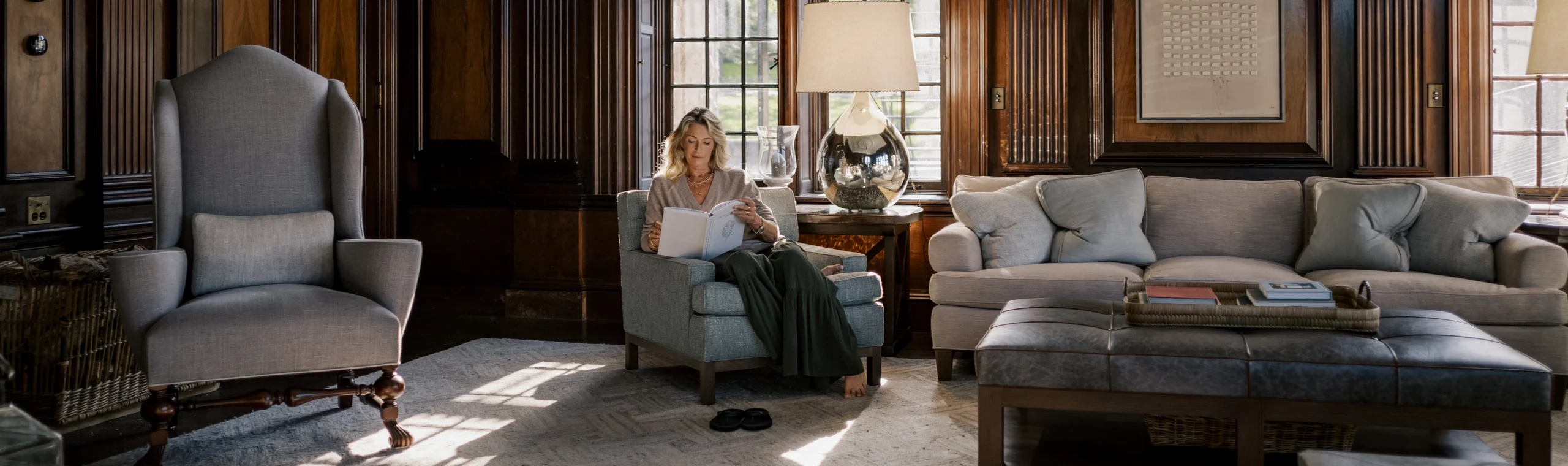 Woman reading a book in a blue armchair in a dark wood-paneled living room with large grid windows and a glass-lamp table nearby.