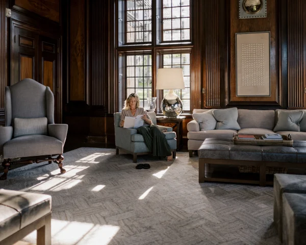 Woman reading a book in a blue armchair in a dark wood-paneled living room with large grid windows and a glass-lamp table nearby.