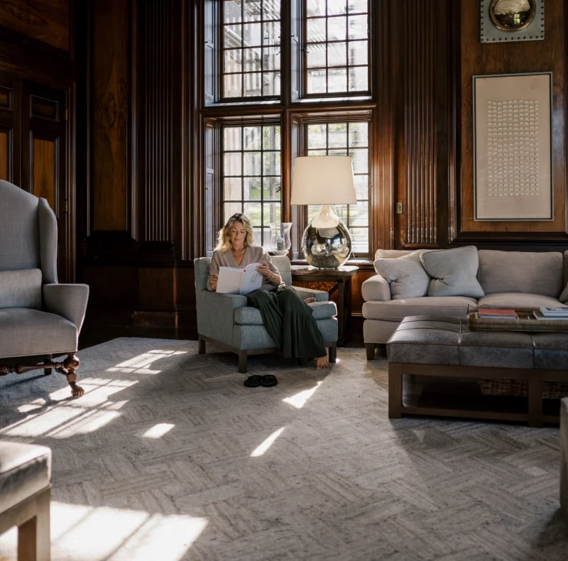 Woman reading a book in a blue armchair in a dark wood-paneled living room with large grid windows and a glass-lamp table nearby.