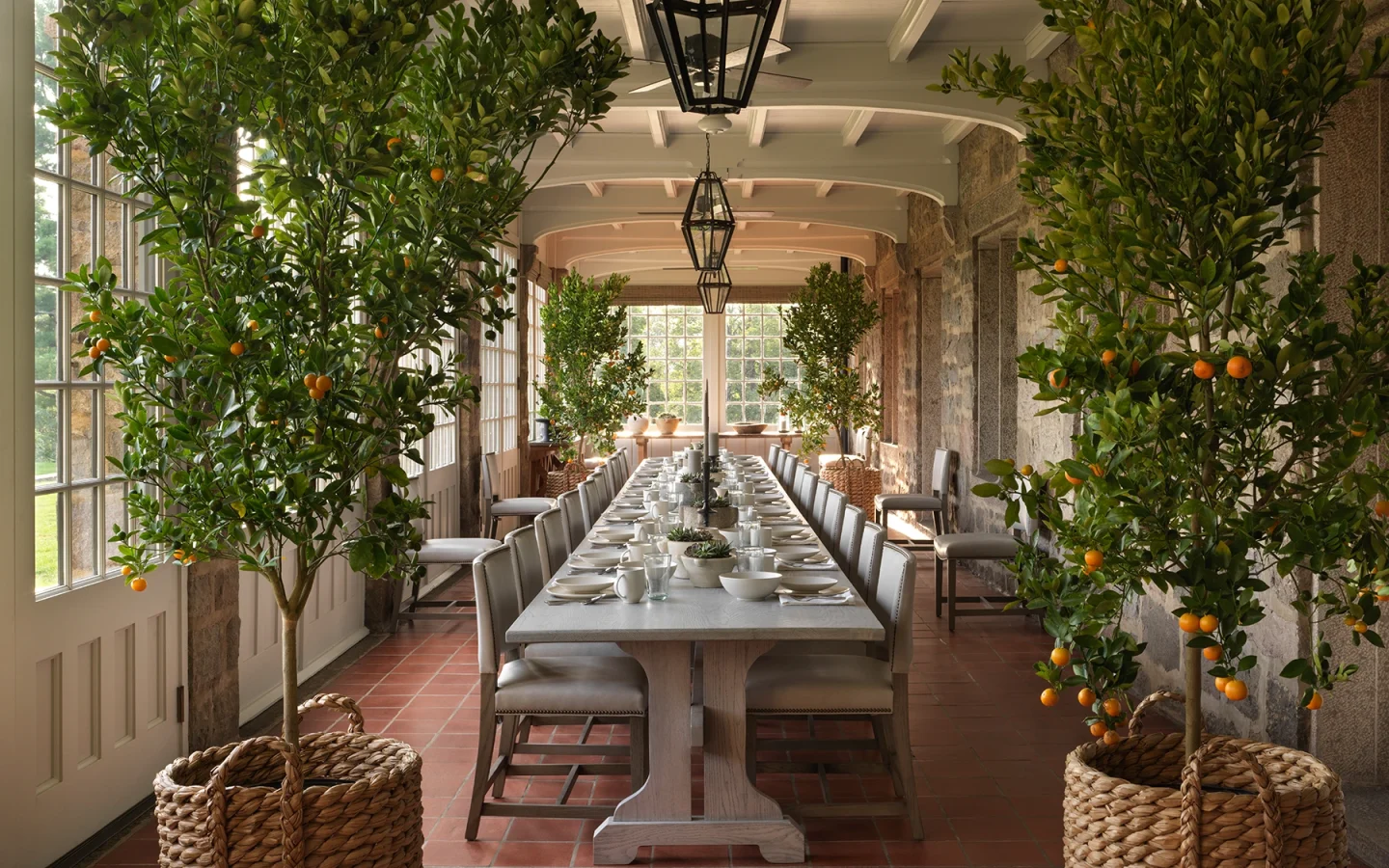 Long dining table in a sunlit conservatory, flanked by orange trees in baskets with place settings and glasses.