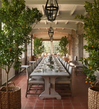 Long dining table in a sunlit conservatory, flanked by orange trees in baskets with place settings and glasses.
