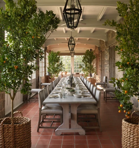 Long dining table in a sunlit conservatory, flanked by orange trees in baskets with place settings and glasses.