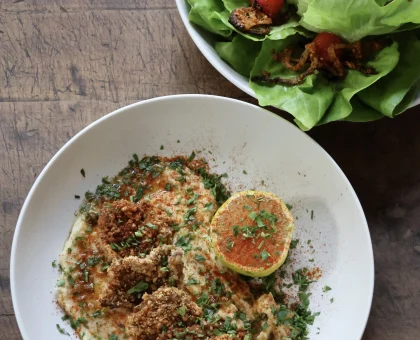 Creamy dish in a white bowl garnished with herbs, crumbled topping, and paprika, with a side salad of greens and cherry tomatoes on a wood table.