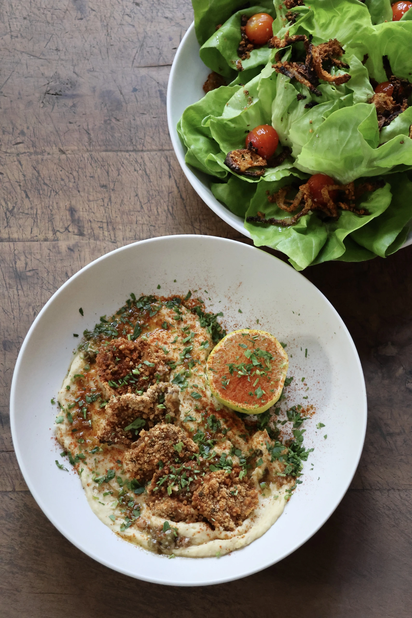Creamy dish in a white bowl garnished with herbs, crumbled topping, and paprika, with a side salad of greens and cherry tomatoes on a wood table.