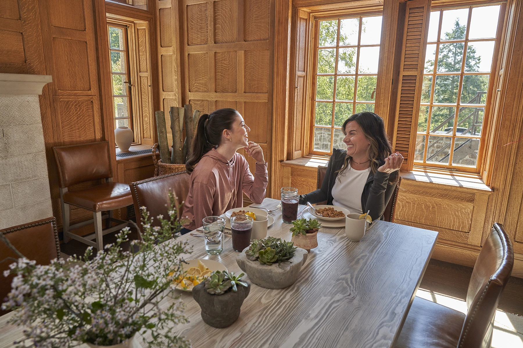 Two women sit across from each other at a sunlit wooden dining table, enjoying breakfast and conversation in a cozy, paneled room with large windows.
