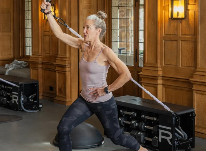 Senior woman in a light pink athletic top and camo leggings lunges on a mat, pulling a resistance rope in a wood-paneled gym.