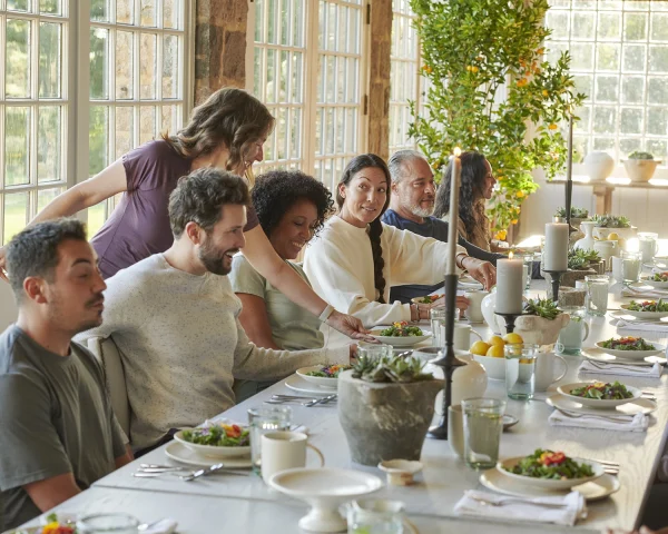 Group of adults sharing a meal around a long, sunlit dining table with salads and candles in a bright room.