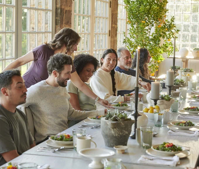 Group of adults sharing a meal around a long, sunlit dining table with salads and candles in a bright room.