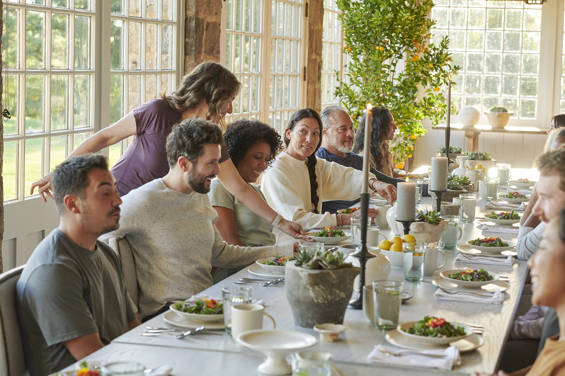 Group of adults sharing a meal around a long, sunlit dining table with salads and candles in a bright room.