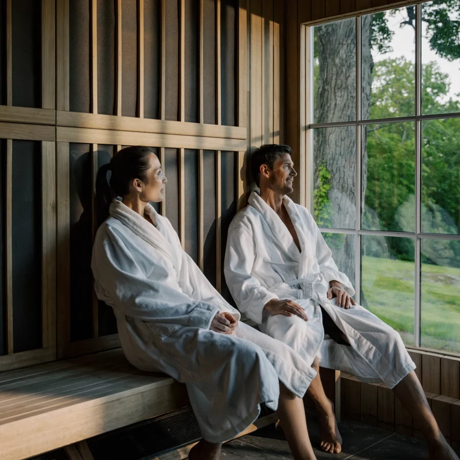 Two people in white bathrobes sit on a wooden bench in a sauna, looking out a large window at green trees outside.