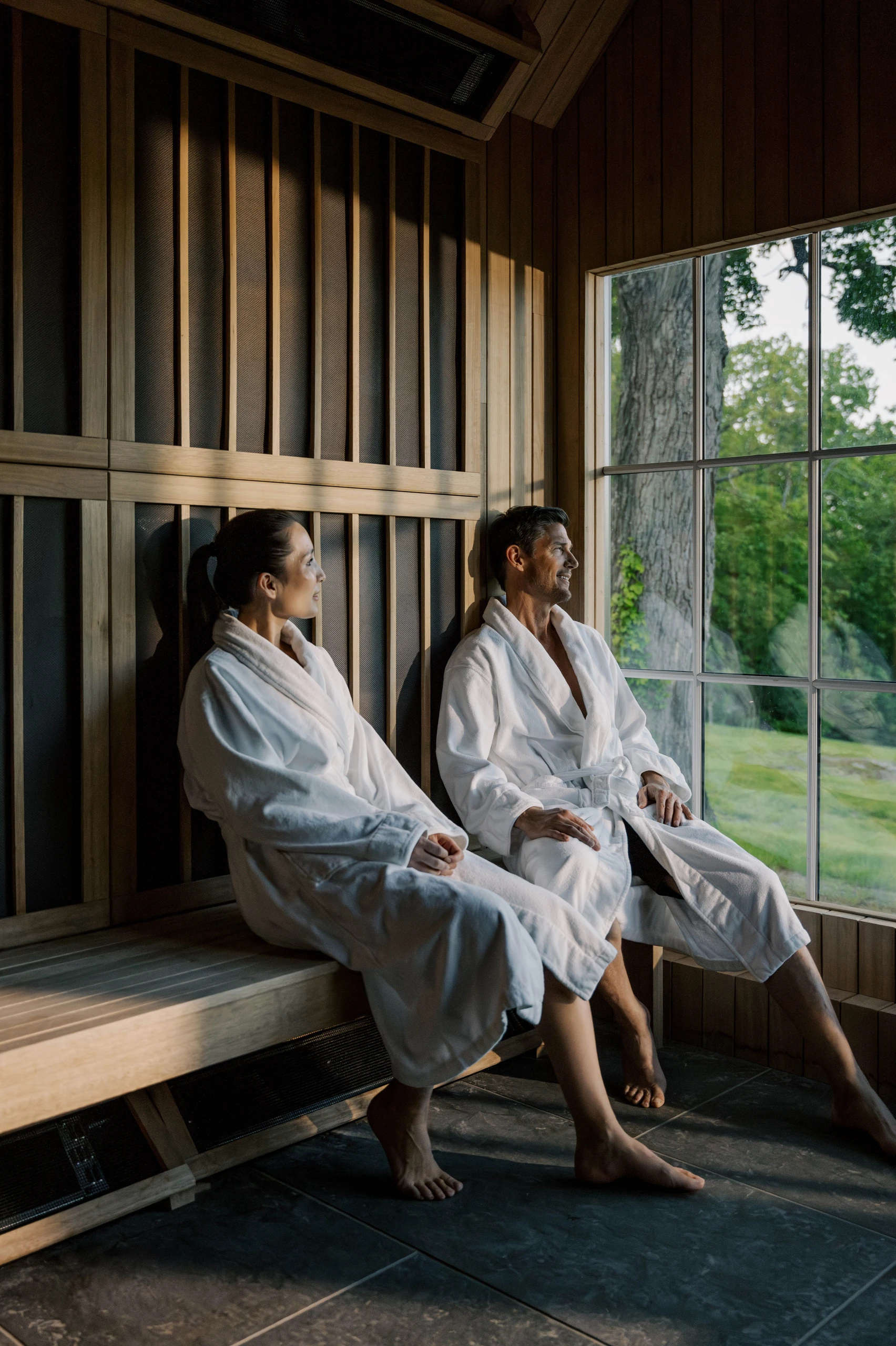 Two people in white bathrobes sit on a wooden bench in a sauna, looking out a large window at green trees outside.