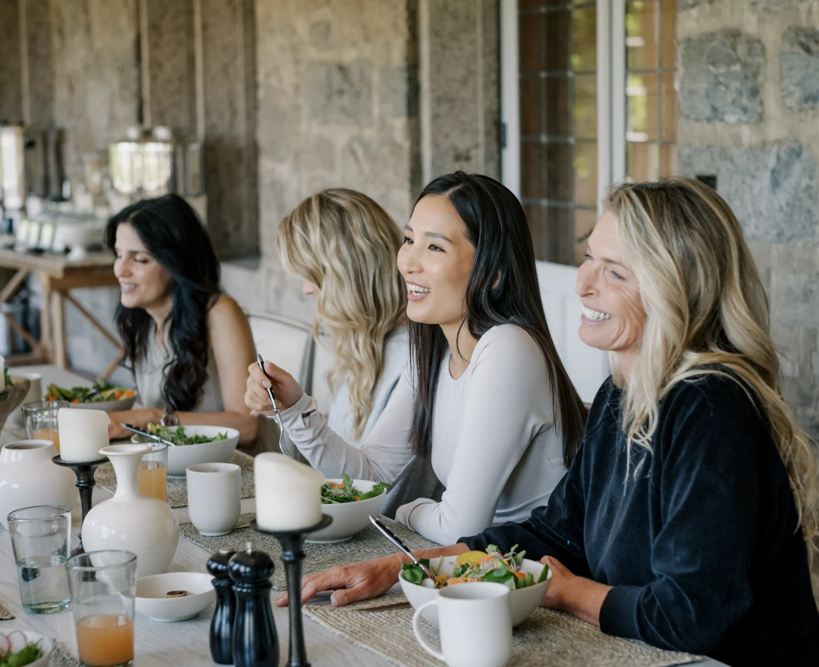 Group of women seated at a long dining table, smiling and eating salad in a stone-walled room.