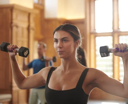 Woman in a black tank top lifting dumbbells in a warm, wood-paneled gym with bright windows behind her.