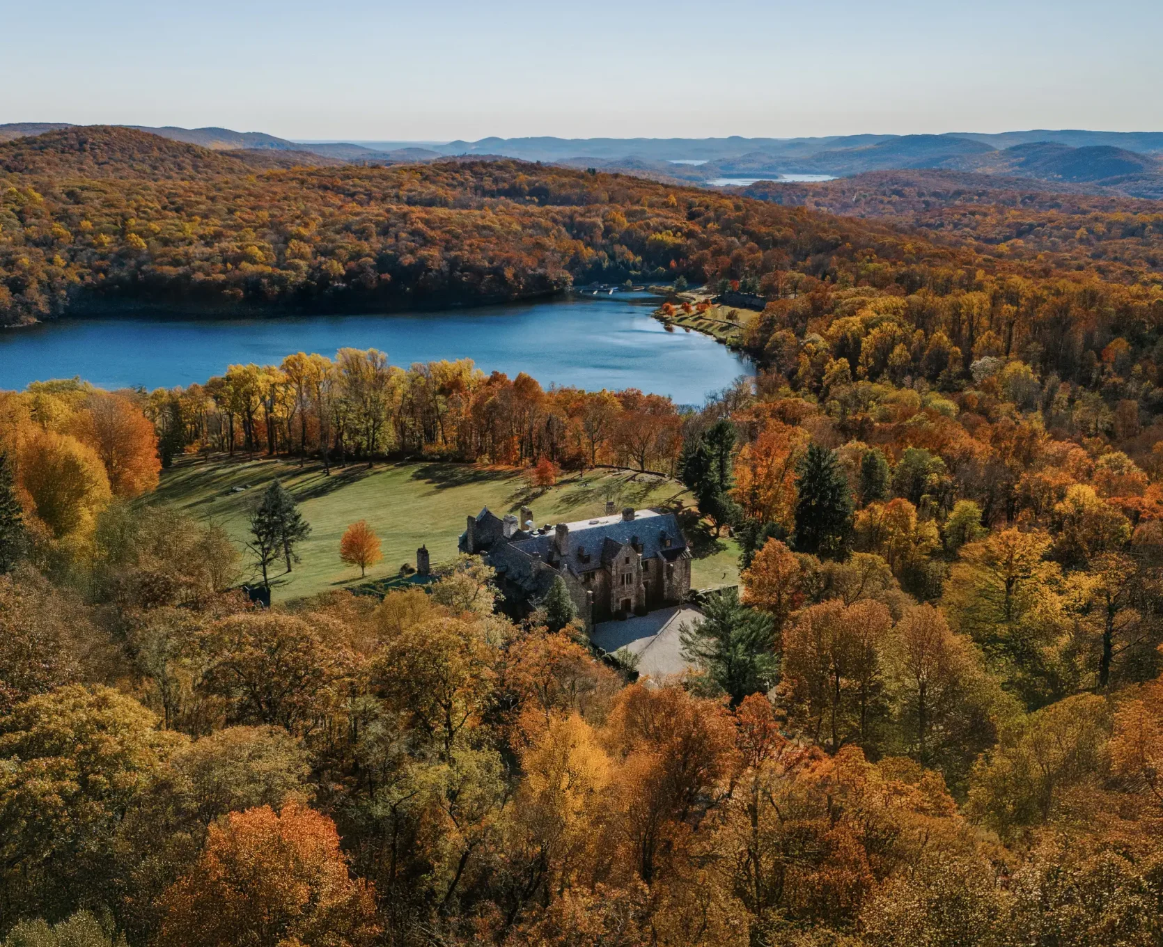 Aerial view of a stone castle amid vibrant autumnal trees, with a blue lake and rolling hills in the distance under a clear sky.