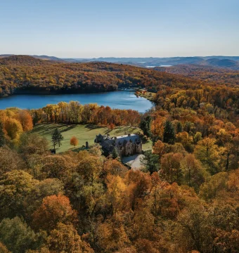 Aerial view of a stone castle amid vibrant autumnal trees, with a blue lake and rolling hills in the distance under a clear sky.