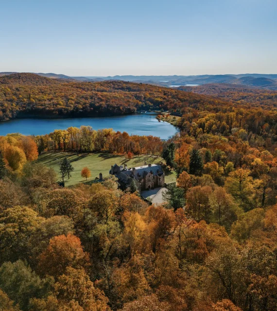 Aerial view of a stone castle amid vibrant autumnal trees, with a blue lake and rolling hills in the distance under a clear sky.